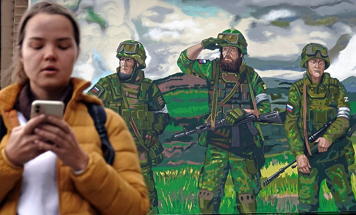 Женщина c мобильным телефоном у фрески, с изображенными на ней солдатами, воюющими в Украине, Москва. Фото: Максим Шипенков / EPA-EFE