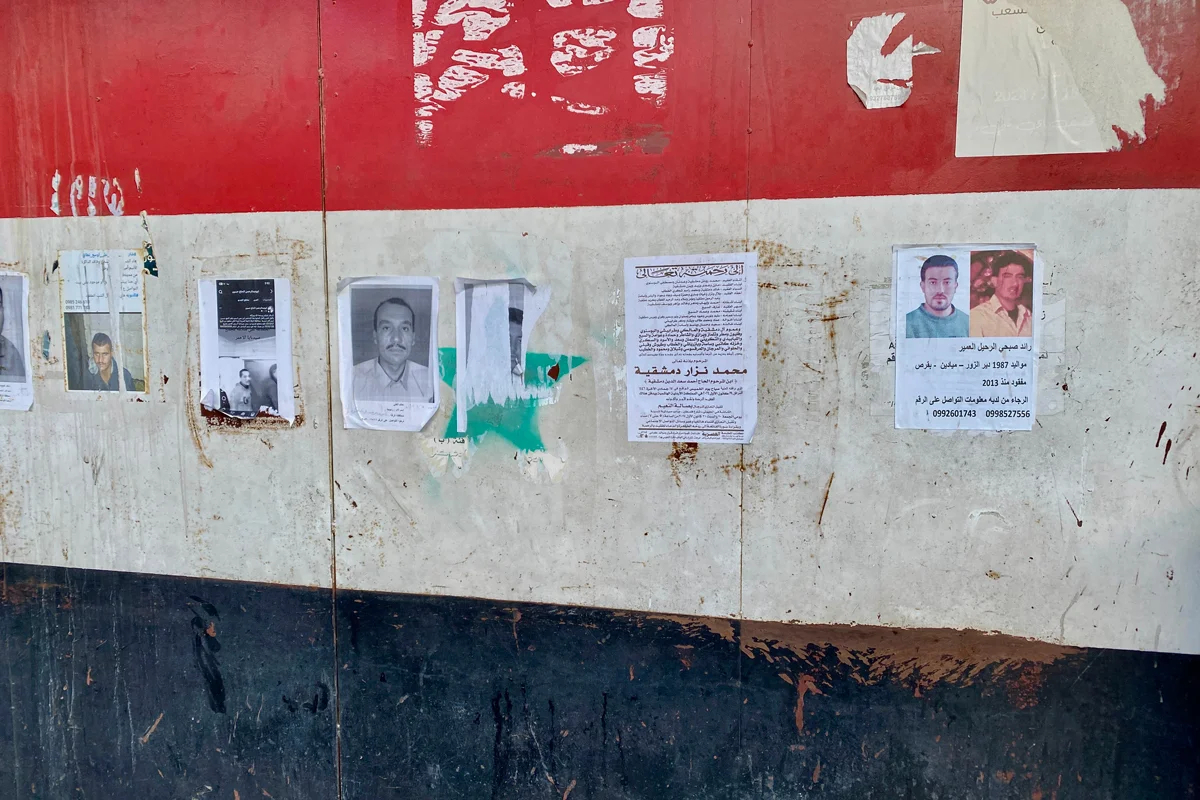 Объявления с фотографиями пропавших людей на стене, выкрашенной в цвета флага Сирии, который использовался при Асаде